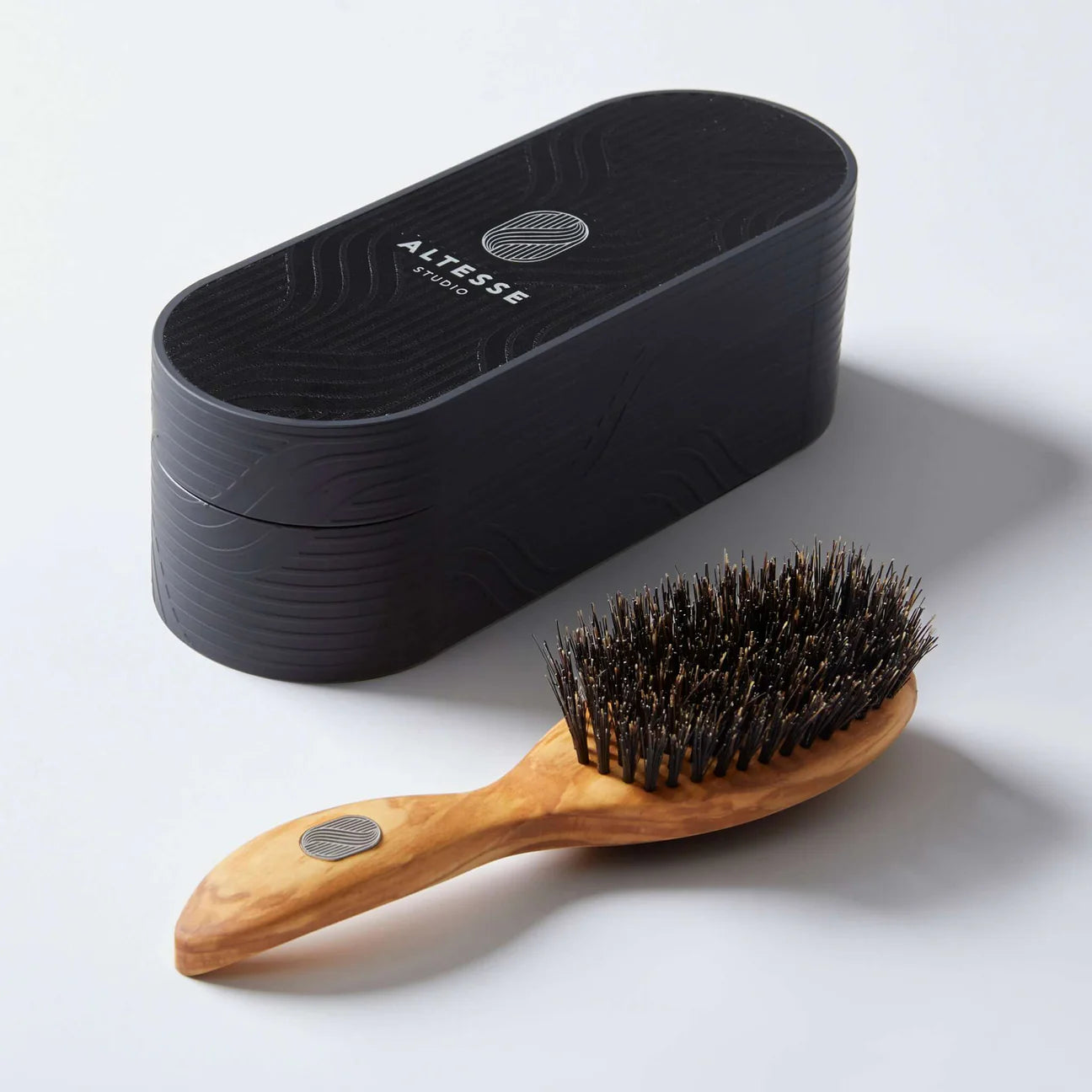 A handmade hairbrush with a wooden handle and boar bristles, accompanied by a black oval-shaped case with the brand name 'Altesse' on it.