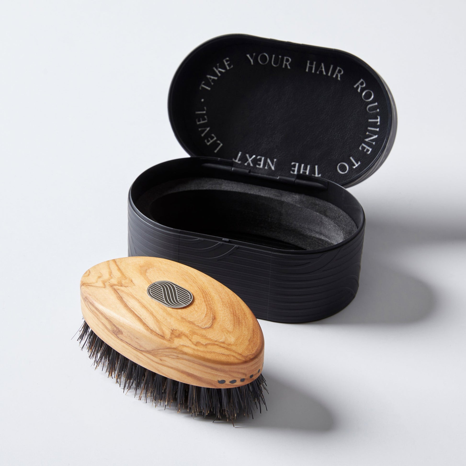 A handmade hair and beard brush with a wooden handle, next to its black leather storage case, with the case lid open.