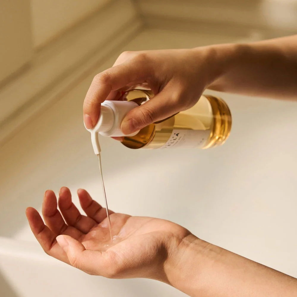 Hand pouring liquid from a gold bottle into another hand against a neutral background