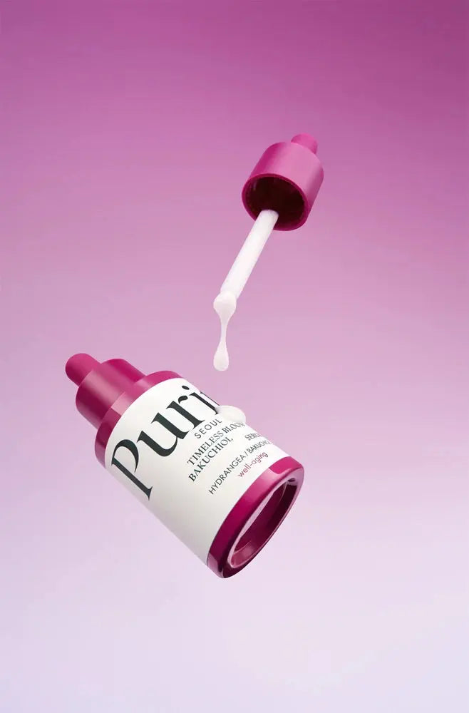 A bottle of Purito Timeless Bloom Bakuchiol serum with its dropper cap removed, showing a small amount of clear liquid serum dripping into the air against a pink background.