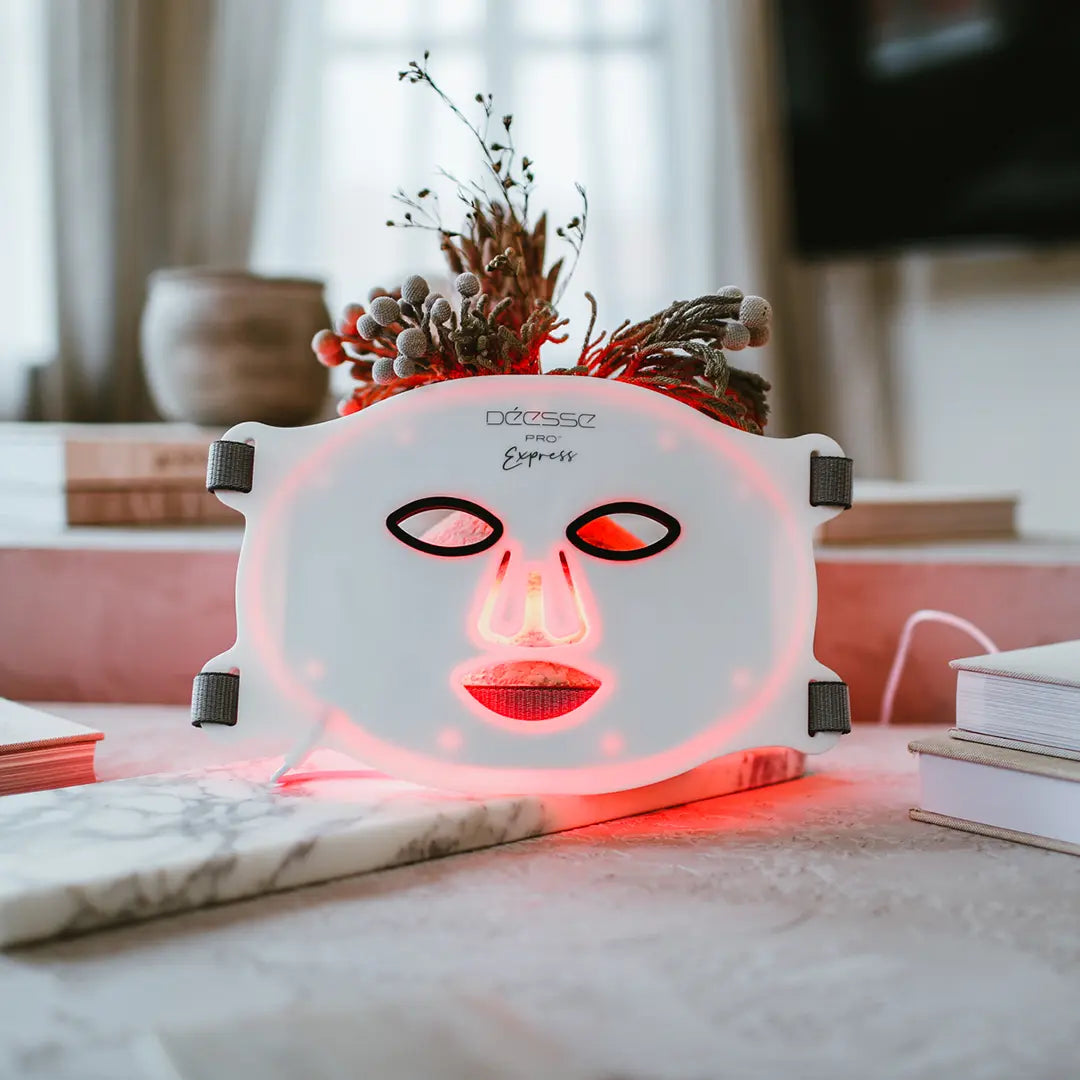 A portable LED light face mask displayed on a flat surface, with red light emitting from the device. The mask appears to cover the face and has various light emitting diodes arranged in a pattern that corresponds to the face's features.